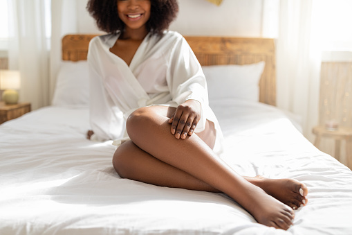 Cropped view of young black woman sitting on bed, wearing white robe, touching her leg, enjoying smooth skin after depilation, indoors. Domestic beauty spa treatment concept