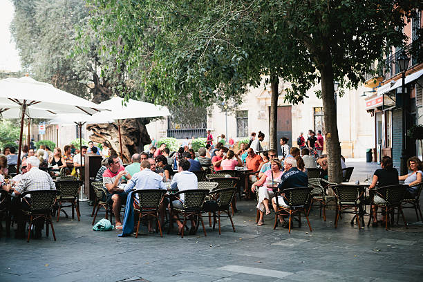Crowd seated outside restaurant in Palma, Majorca This long shot shows many diverse people sitting in an outdoor dinning area near a restaurant in Palma, Majorca.  The tables are covered by tall, white, fabric, sun-shade umbrellas on the left and mature, dark green trees on the right.  The outdoor restaurant has charcoal gray pavement in front and old buildings in the background. patio stock pictures, royalty-free photos & images