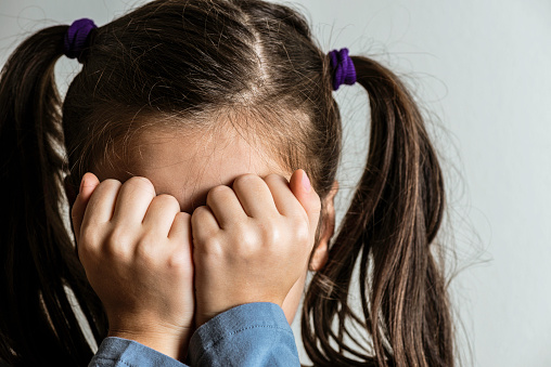 Little crying caucasian girl with double ponytail who is hiding her face with her hands. Representing child abuse and domestic violence.