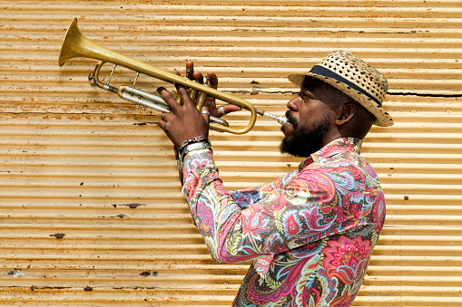 A well known professional Cuban musician with a beard, wearing a Panama hat, standing against a closed yellow store shutter, playing a trumpet outdoors, Havana, Cuba, 50 megapixel image