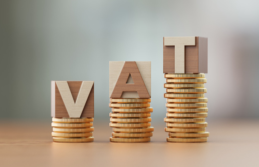 Cube blocks made of wood material are standing on coin towers to form VAT text over defocused background. Horizontal composition with copy space. Great use for finance concepts.