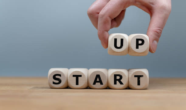Cubes form the words "START UP" while to fingers lift the letters "UP" in the air. Cubes are on a wooden table in-front a grey background. stock photo