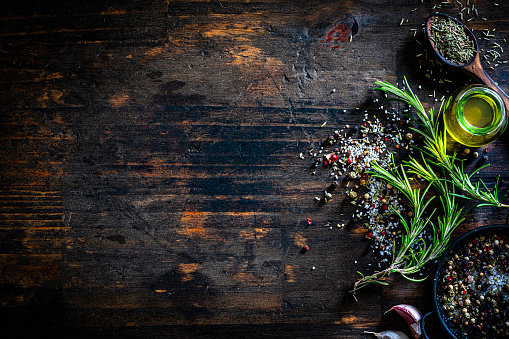 Culinary background: overhead view of rosemary twigs, marine salt, pepper, olive oil container arranged at the right of a dark wooden table making a border and leaving useful copy space for text and/or logo. Predominant colors are dark brown and green. High resolution 42Mp studio digital capture taken with Sony A7rII and Sony FE 90mm f2.8 macro G OSS lens