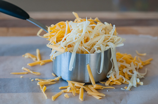 Close-up of overflowing measuring cup of grated orange and white cheese. Jack and cheddar cheese. Pile of grated cheese in a stainless steel measuring cup.