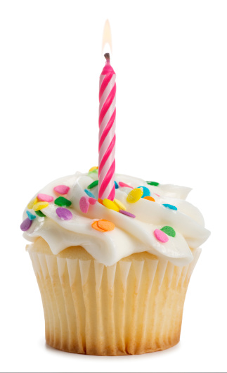 This is a photo of a vanilla cupcake with a lit pink candle, icing and sprinkles on top. This cupcake is isolated on a white background with a drop shadow. 