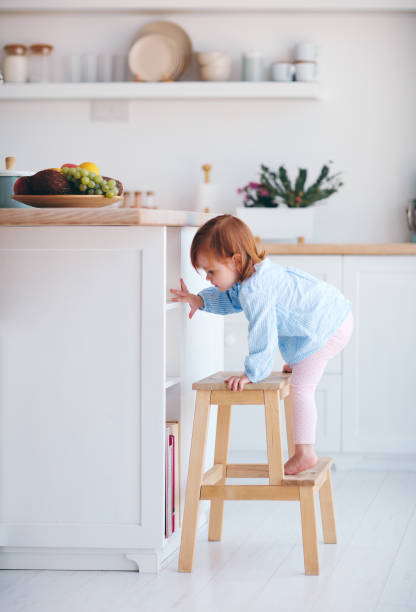 curious infant baby girl trying to reach the fruit on the table in the kitchen with the help of step stool stock photo