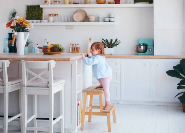 curious infant baby girl trying to reach things on the table in the kitchen with the help of step stool stock photo