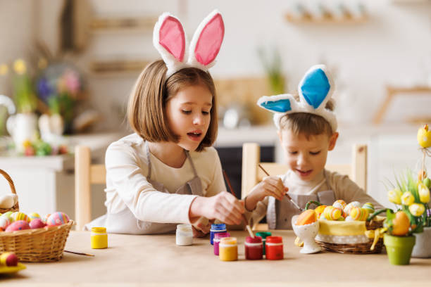 Curious kids brother and sister in bunny ears painting Easter eggs together in kitchen Curious kids brother and sister in plush bunny ears preparing for Easter together, children painting  eggs with   paints and brushes while sitting at kitchen table. Spring holidays concept easter sunday stock pictures, royalty-free photos & images