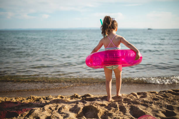 Curious little girl stepping into sea with a pink inflatable ring and scuba mask Excited girl with a scuba mask and inflatable ring having fun at the beach holiday little girls in bathing suits stock pictures, royalty-free photos & images