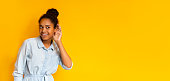 Curious snoopy schoolgirl holding hand at her ear, trying to overhear private conversation, posing at yellow studio background. Panorama with free space