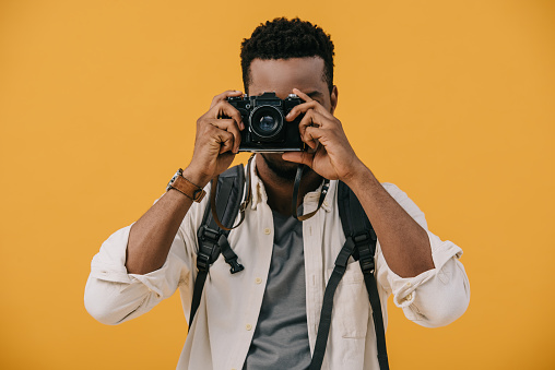 curly african american photographer covering face with digital camera isolated on orange