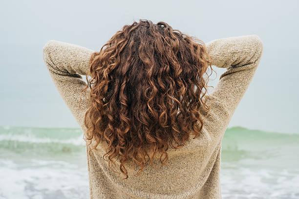 Curly brunette in a sweater on the coast Curly brunette in a sweater on the coast faces the sea and straightens hair, view from the back curly hair stock pictures, royalty-free photos & images