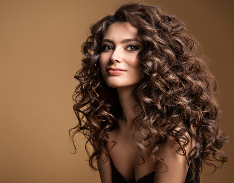 Curly Hair Model. Woman Wavy Long Hairstyle. Brunette Fashion Girl with Volume Hairdo and Natural Make up over Beige Studio Background