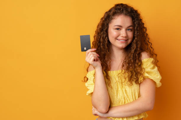 Curly-haired woman holding black credit card against yellow background stock photo