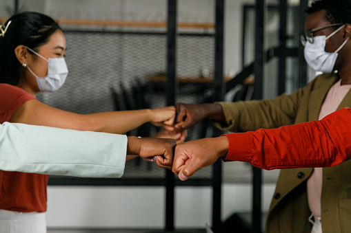 Side view of unrecognizable group of four diverse business people standing opposite each other and fist bumping when greeting after a meeting. They are wearing protective face masks.