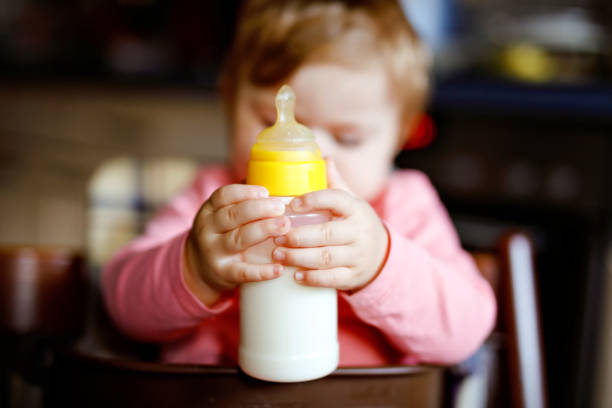 urocza urocza dziewczynka trzymająca butelkę pielęgniarską i mleko z formuły do picia. pierwsze jedzenie dla niemowląt. nowo narodzone dziecko, siedzące na krześle kuchni domowej. zdrowe dzieci i koncepcja karmienia butelką - baby formula zdjęcia i obrazy z banku zdjęć