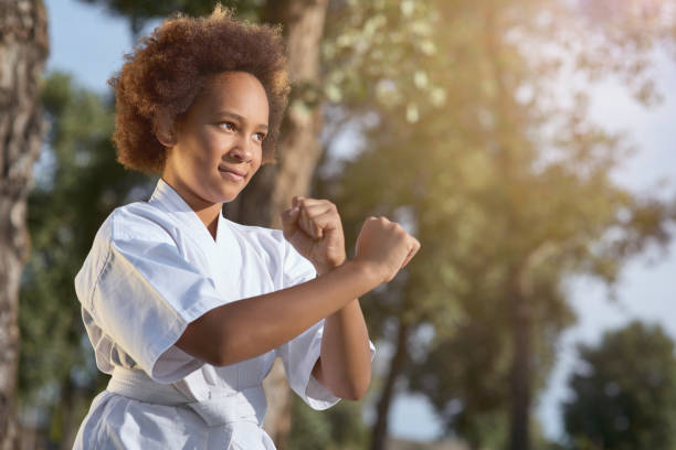 Cute Afro American girl practicing martial arts on sunny day in park Adorable female child karateka looking away and smiling while doing fighting stance on the street "martial arts" stock pictures, royalty-free photos & images
