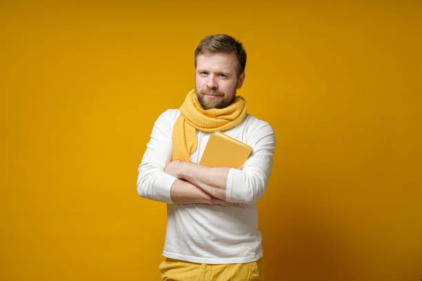 Cute bearded student in a yellow knitted scarf holds a book in his crossed arms and looks confidently. Education concept. stock photo