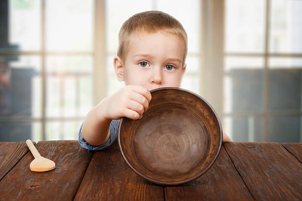 Cute blonde boy shows empty plate, hunger concept Child hunger concept. Small toddler boy shows empty bowl sitting at dark wood table with wooden spoon. Cute child has no food in plate hungry stock pictures, royalty-free photos & images