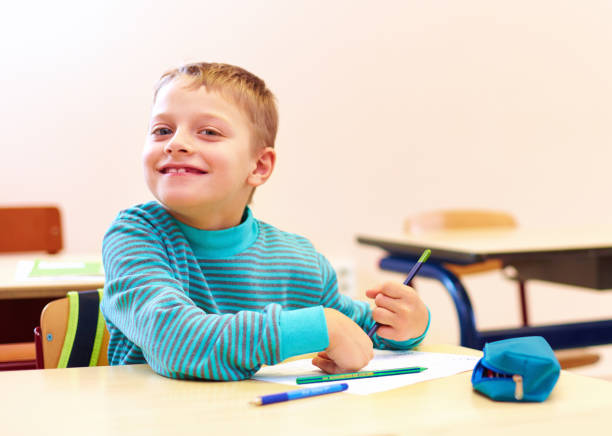 cute boy with special needs writing letters while sitting at the desk in class room stock photo