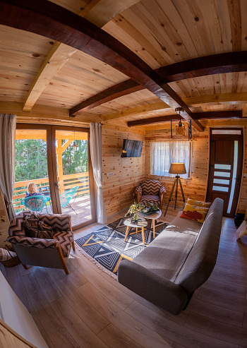 The interior design of a living room with large windows in a tiny rustic log cabin. An unrecognizable woman is sitting on the porch