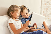 We love internet. Two happy kids looking at pad screen and using headphones while sitting on bed together