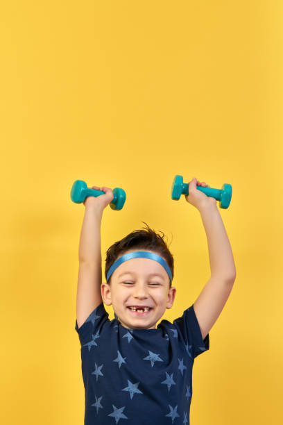 Cute child is lifting dumbbell for healthy lifestyle on yellow background. He is happy and proud.