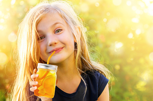 Cute funny little girl drinking mango orange beverage through a strawi in mason jar in hot Summer day on nature background