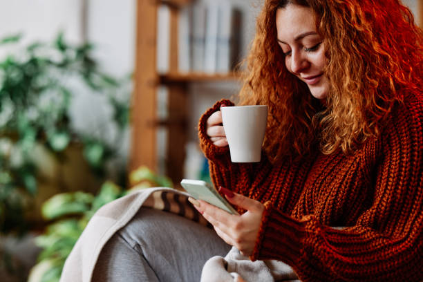 A cute ginger girl with curly hair is sitting in the chair at home in the morning, texting messages on the phone and drinking her coffee. stock photo