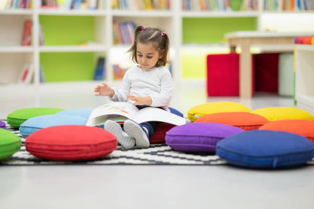 Cute girl is reading book sitting in nursery library Cute girl is reading book sitting in nursery library indoor playground stock pictures, royalty-free photos & images