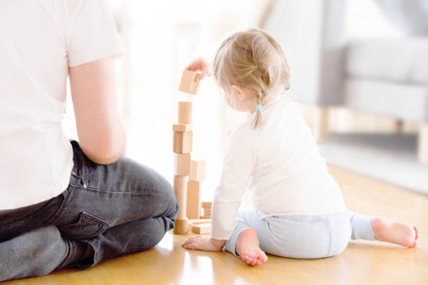 Cute girl playing with wooden blocks ont he floor with her father Cute two year old girl is sitting on the floor playing with wooden blocks at home with her father. The photo is taken from behind them and you can see the tower of blocks they have built between them. A typical family moment at home. swedish girl stock pictures, royalty-free photos & images