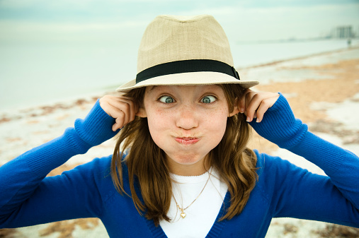 A cute young girl with a panama hat is making a funny face on the beach. She is wearing a royal blue cardigan on a white t-shirt. Hair is mid-long and eyes very blue. This was taken in Clearwater, Florida, Shallow DOP, focus on the face. Horizontal with copy space.