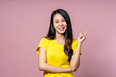 黄色のカジュアルシャツを着たかわいい幸せなアジアの女性は、カメラを見て、テキストメッセージや広告のためのピンクのスタジオの背景に笑顔と指を指しています。