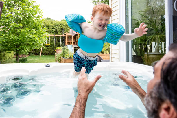Cute Kid and Father in the Hot Tub in the Backyard in Summer A little redhead boy is having fun in the hot tub in his backyard in summer. He is jumping in his father’s arms. The cute kid is laughing and having fun. He is wearing a life jacket for safety. Their swimming pool is a spa. hot tub stock pictures, royalty-free photos & images