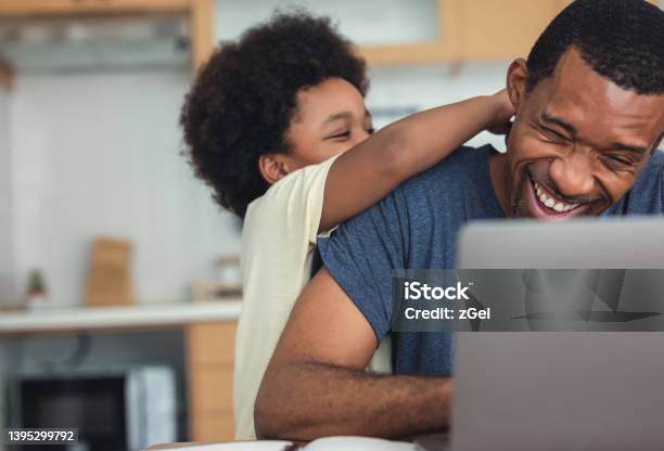 Cute little African American boy hugging and playing with dad while his dad working from home with laptop computer for remoter job in modern kitchen. Concepts of remote work, children, parenthood