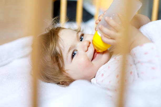 formül hafif şişe tutan ve içme sevimli küçük bebek kız. çocuk uykudan önce bebek karyolası yatakta - baby formula stok fotoğraflar ve resimler