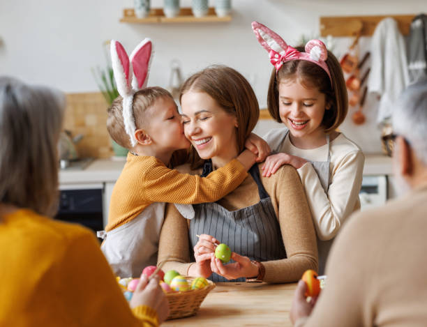 Cute little children embracing and kissing young happy mother while painting Easter eggs with family stock photo