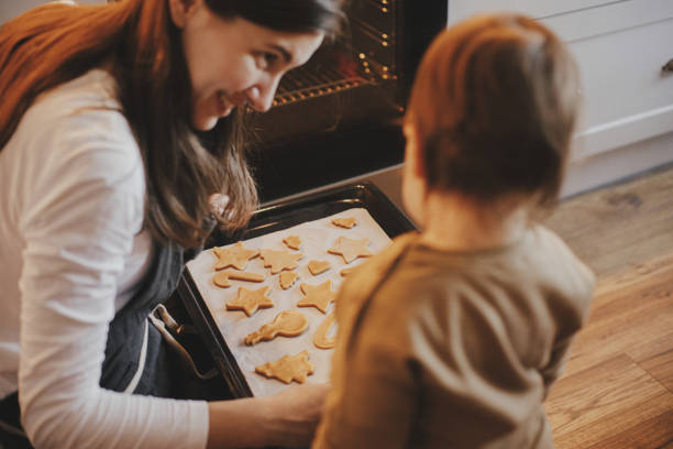 Cute little daughter and mother baking christmas gingerbread cookies in modern scandinavian kitchen. Cute toddler girl and mom putting tray with cookies in oven. Family holiday preparations stock photo