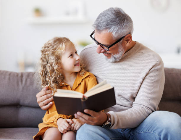 Cute little girl granddaughter reading book with positive senior grandfather stock photo