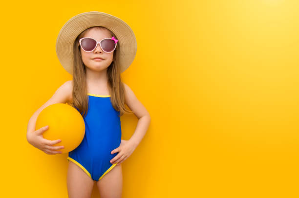 Cute little girl in a blue swimsuit for a mockup on a yellow background. Beautiful fashionable child in a hat and sunglasses is holding a ball. Summer vacation concept. Place for text Cute little girl in a blue swimsuit for a mockup on a yellow background. Beautiful fashionable child in a hat and sunglasses is holding a ball. Summer vacation concept. Copyspace little girls in bathing suits stock pictures, royalty-free photos & images