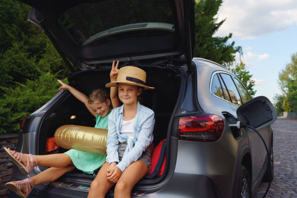 Cute little sisters sitting in trunk while waiting for charging car before travelling on summer holiday. stock photo