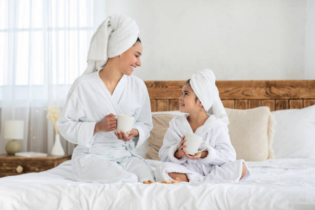 Cute mother and daughter drinking tea in bed Cheerful cute young mother and little daughter drinking tea in bed, wearing white bathrobes and having towels over their heads, enjoying beauty day together at home, copy space hot middle eastern girls stock pictures, royalty-free photos & images