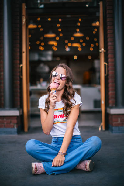 Cute Portrait of a Beautiful Young Woman Sitting and Enjoying a Pink Ice Cream Cone Cute Portrait of a Beautiful, Fashionable Young Woman Sitting Cross-Legged and Enjoying a Pink Ice Cream Cone with Pretty Bokeh Lights in the Background in a Colorado Mountain Town skinny food co stock pictures, royalty-free photos & images