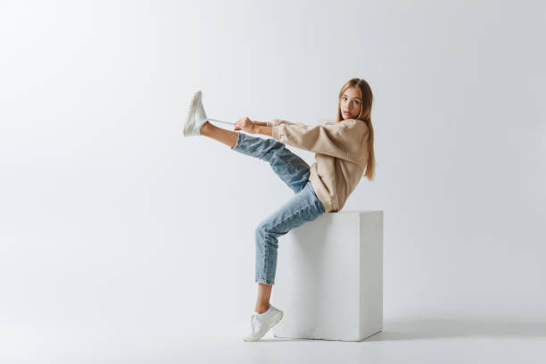 Cute teenage girl lacing her sneakers, pulling on laces. Sitting on a white cube stock photo