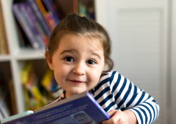 Cute toddler holds a storybook from her bookcase to read stock photo