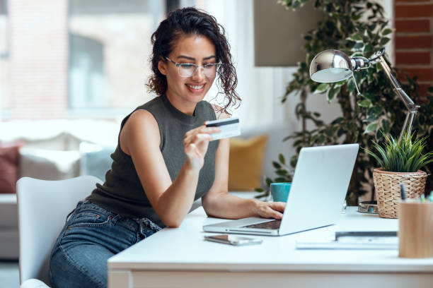 Cute woman holding white credit card for shopping online with computer while sitting in living room at home. stock photo