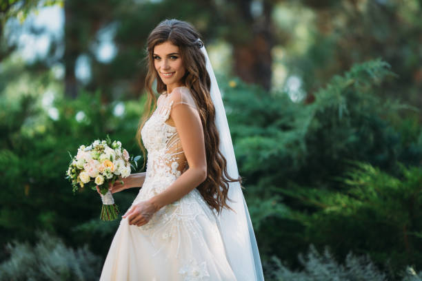 Cute young bride with long hairs holding her wedding bouquet includes white roses and other flowers. Beautiful white marriage dress. Pretty girl on greet trees background stock photo