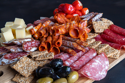 Cutting board with ham, salami, cheese, crackers and olives on a dark stone background.