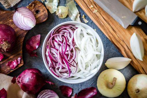 Top view of a bowl full of red and golden sliced onions surrounded by a wooden cutting board with a kitchen knife on top and whole and sliced golden and red onions. The bowl is at the center of the image. Low key DSLR photo taken with Canon EOS 6D Mark II and Canon EF 24-105 mm f/4L