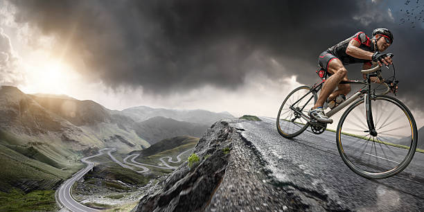 Cyclist Climbs To The Top Wide angle close up image of a cyclist riding on a the peak of a road winding through mountains in a generic location, under a dramatic stormy evening sky at sunset. The bike is generic and cyclist is wearing an unbranded cycling suit, helmet and sunglasses. With intentional lighting effects.  extreme sports stock pictures, royalty-free photos & images